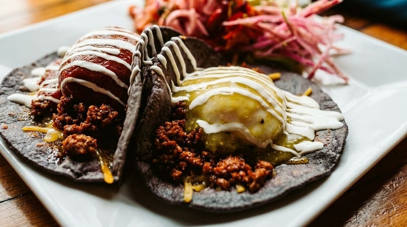 Two blue corn tacos with chorizo, one topped with red sauce, the other with green, drizzled with crema.