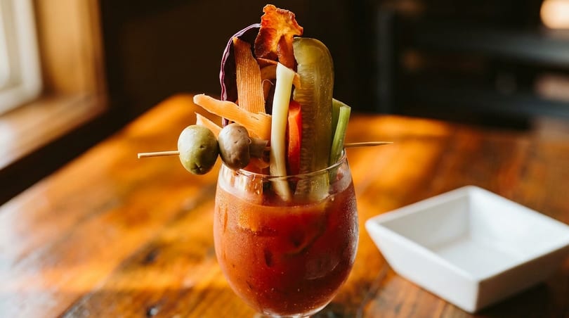 A Bloody Mary cocktail overflowing with olives, pickles, celery, and other garnishes on a wooden table.