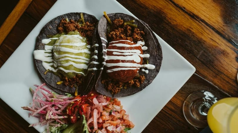 Two dark tacos with green and red sauces, garnished with pico de gallo and radishes.