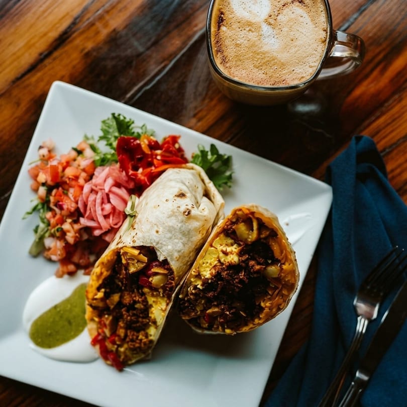 A breakfast burrito, cut in half, with salsa, pickled onions, and a latte on a wooden table.