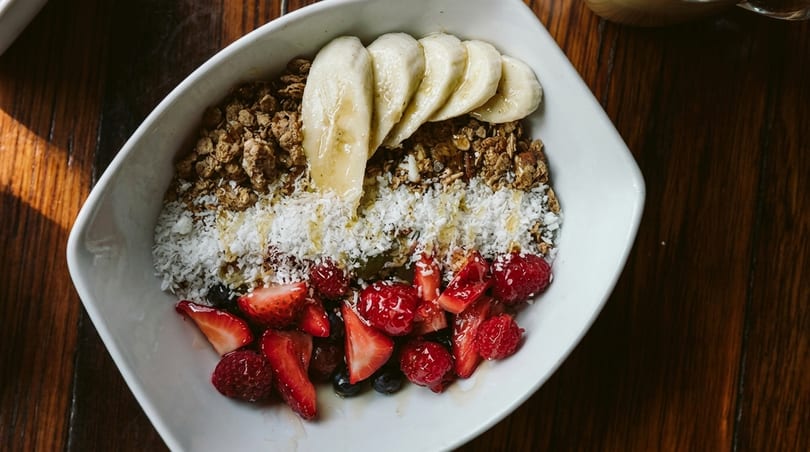 Overhead view of a breakfast bowl with granola, banana slices, shredded coconut, strawberries, and raspberries.