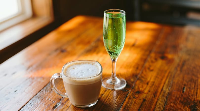 A latte in a clear mug and a green sparkling drink in a champagne flute on a wooden table.
