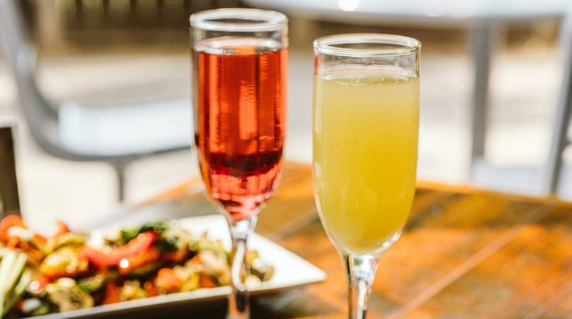Two flutes of sparkling drinks, one red and one yellow, with a plate of food in the background.