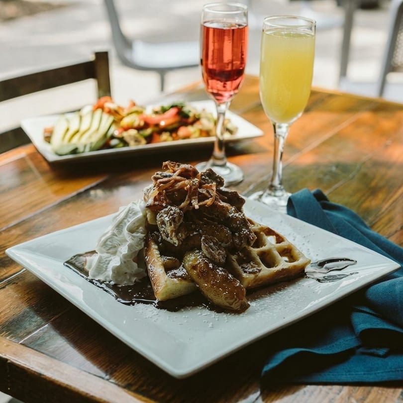 Waffle with whipped cream, caramelized bananas, and fried onions, with mimosas and avocado toast in background.