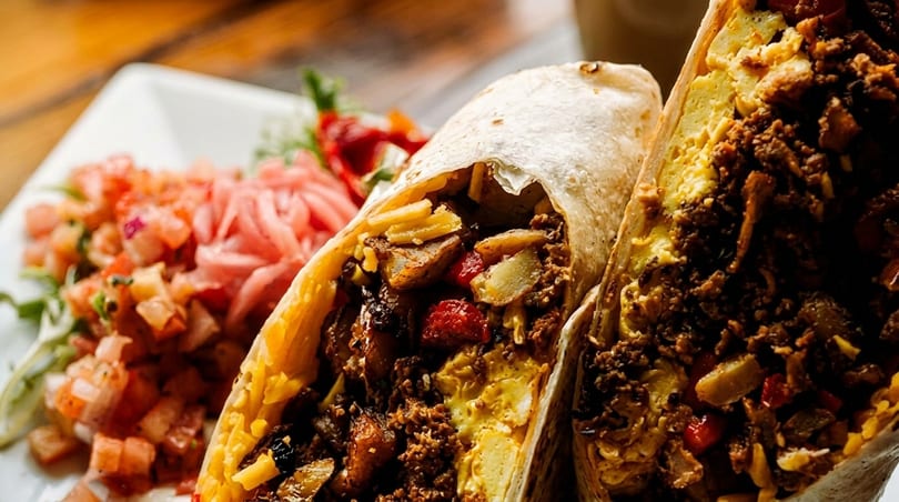 Two breakfast burritos filled with eggs, meat, potatoes, and peppers, served with salsa.