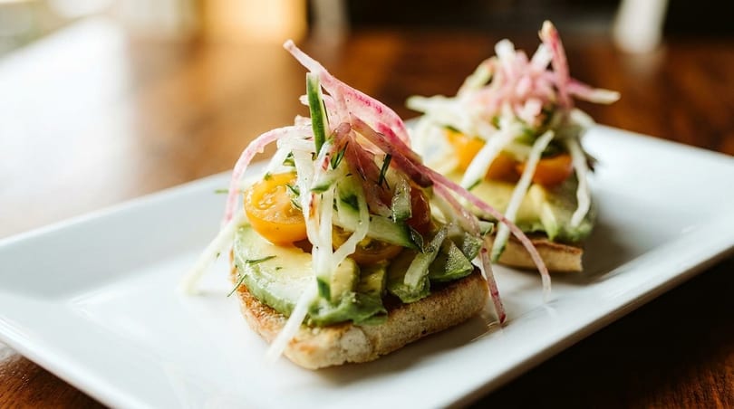 Two avocado toasts topped with cherry tomatoes, cucumber, and pickled red onions on a white plate.