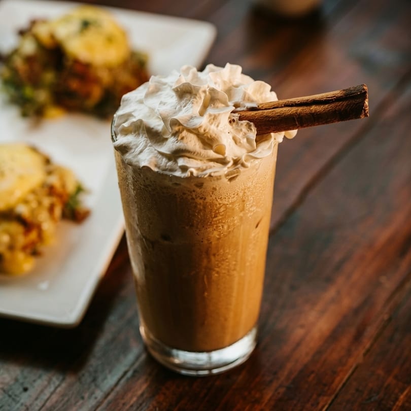 Iced coffee with whipped cream and a cinnamon stick, with food blurred in the background.