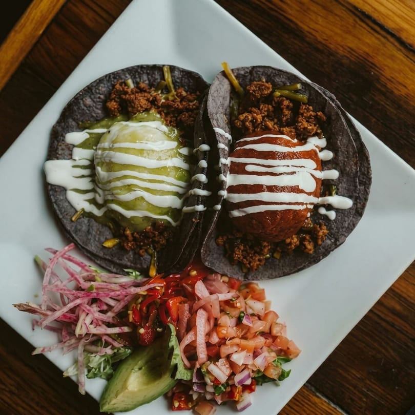 Two dark tacos with meat, guacamole, and sour cream, served with salsa and avocado.