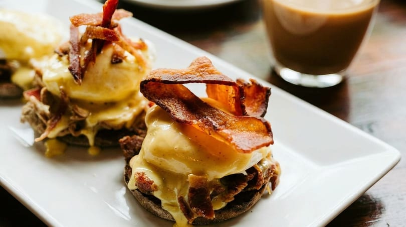 Two eggs benedict with hollandaise sauce and crispy bacon on a white plate with coffee.